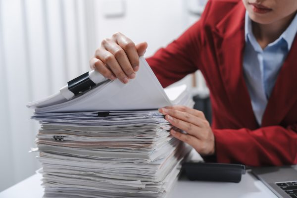 close up hands of person in a red blazer organizing a substantial stack of various papers, likely sorting or reviewing documents for yearly tax purposes.