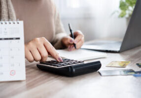 woman calculating monthly debt payments, writing down numbers with a calendar with marked due dates, using a calculator with credit card on the desk