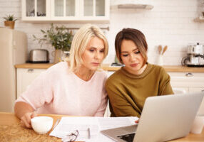 serious dark haired girl sitting at kitchen table with her middle aged mother using wifi on generic portrable computer, drinking coffee and calculating family budget, planning expensive purchase