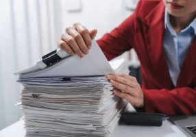 close up hands of person in a red blazer organizing a substantial stack of various papers, likely sorting or reviewing documents for yearly tax purposes.